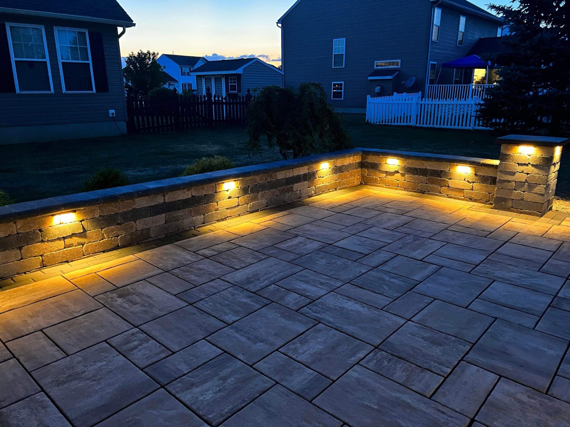 A patio with illuminated brick wall lights during twilight. Houses with closed windows surround the area, creating a cozy suburban atmosphere.