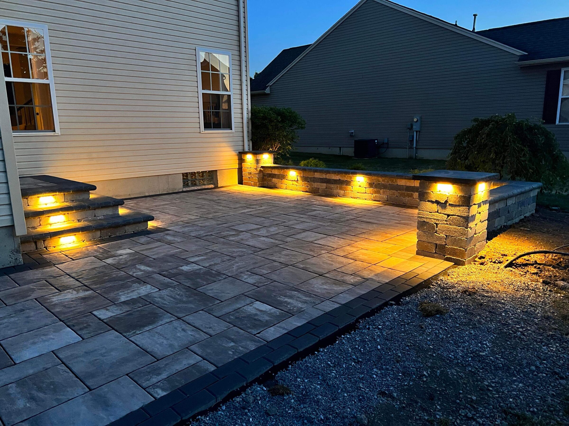 A well-lit stone patio with steps and retaining walls, adjacent to a beige house. Evening ambiance, creating a cozy outdoor space.