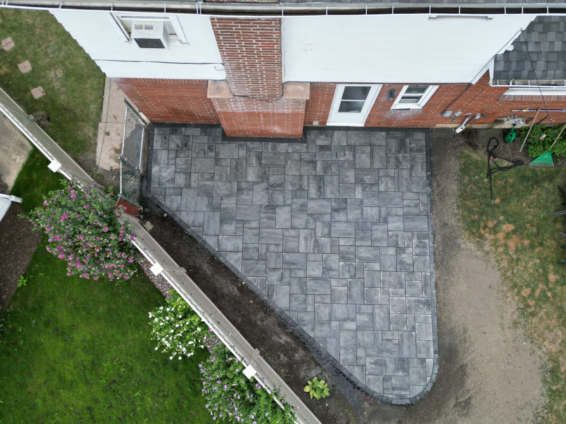 Aerial view of a brick house with a newly paved stone patio, bordered by grass and flowers in a small backyard space.