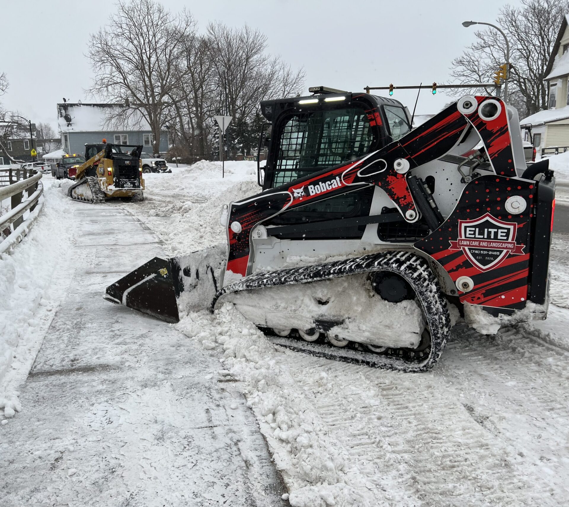 Snow plows clear a snowy sidewalk, while bare trees line the road and a traffic light is visible in the background.