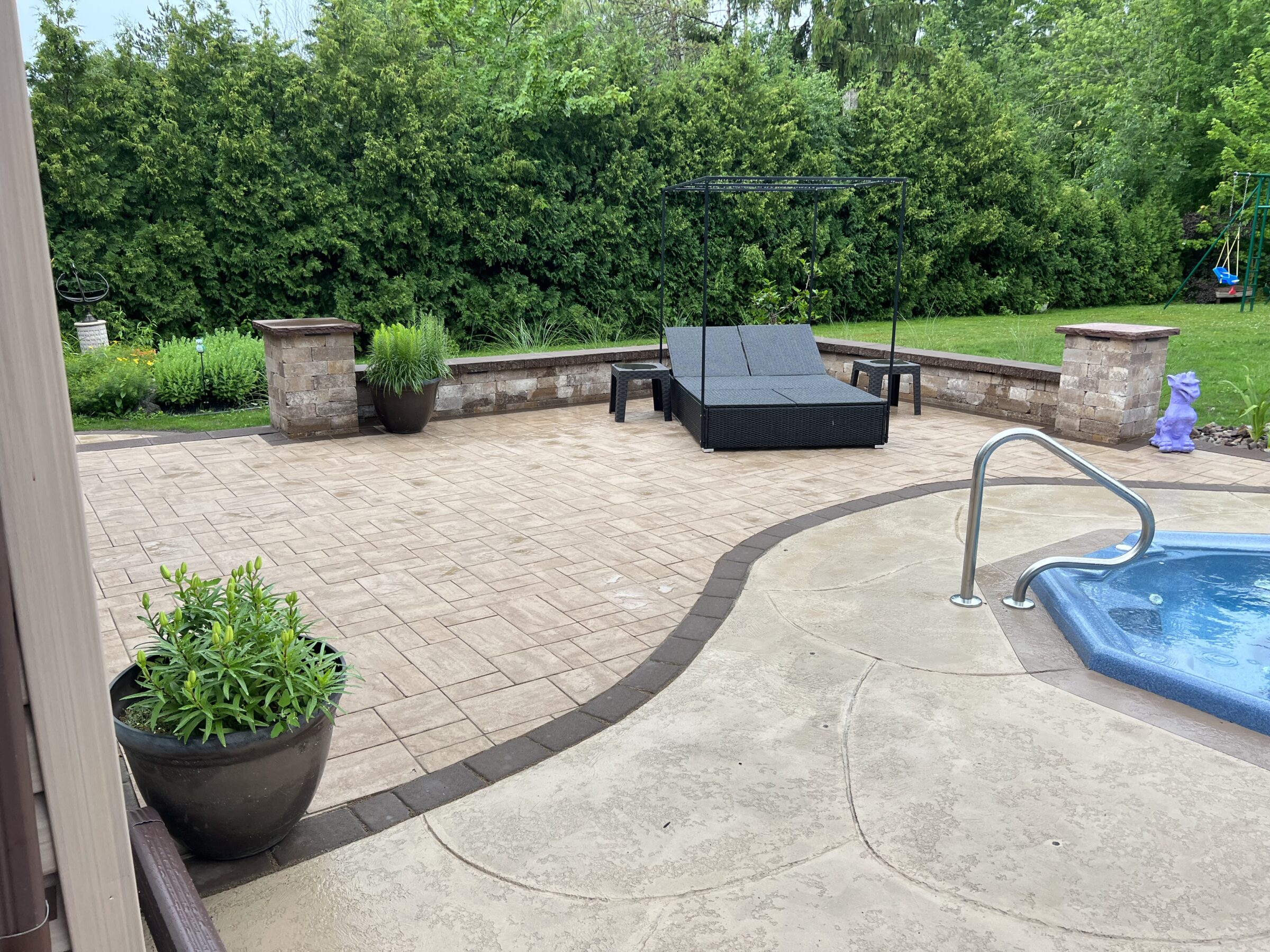 A backyard patio with lounge chairs, a swimming pool, and lush greenery. Potted plants add decoration to the paved area.