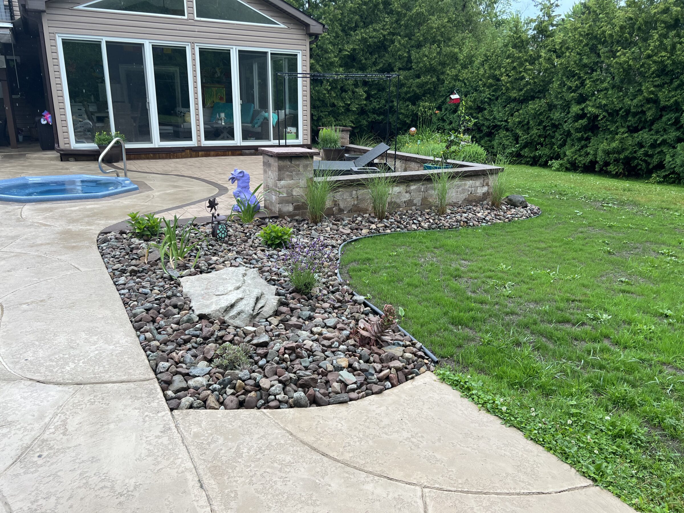 Backyard garden with a small rock and plant bed beside a swimming pool. Sliding glass doors lead to the patio. Lush greenery surrounds.
