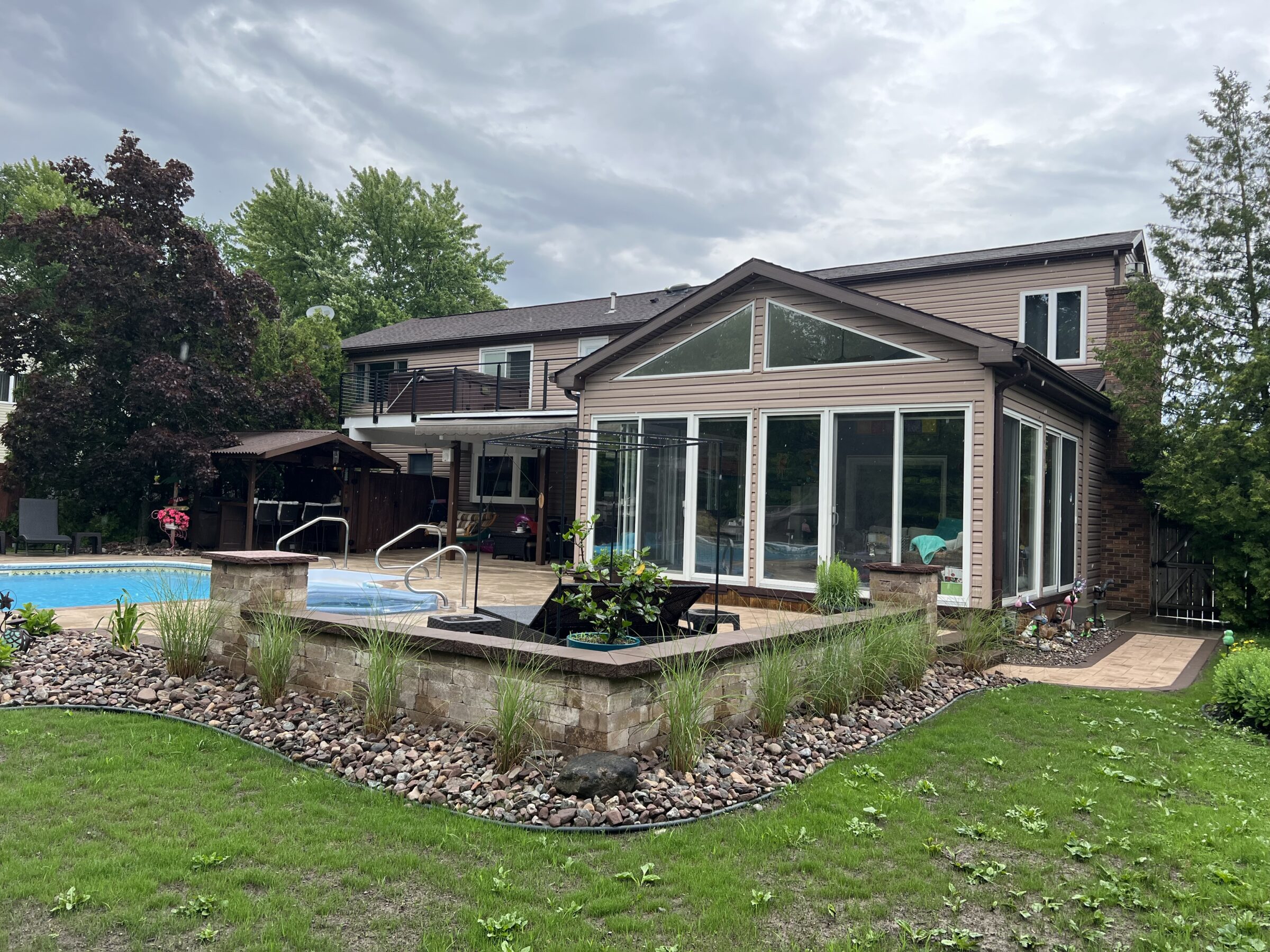 A two-story house with a modern sunroom, outdoor pool, and landscaped garden under a cloudy sky, surrounded by lush greenery.
