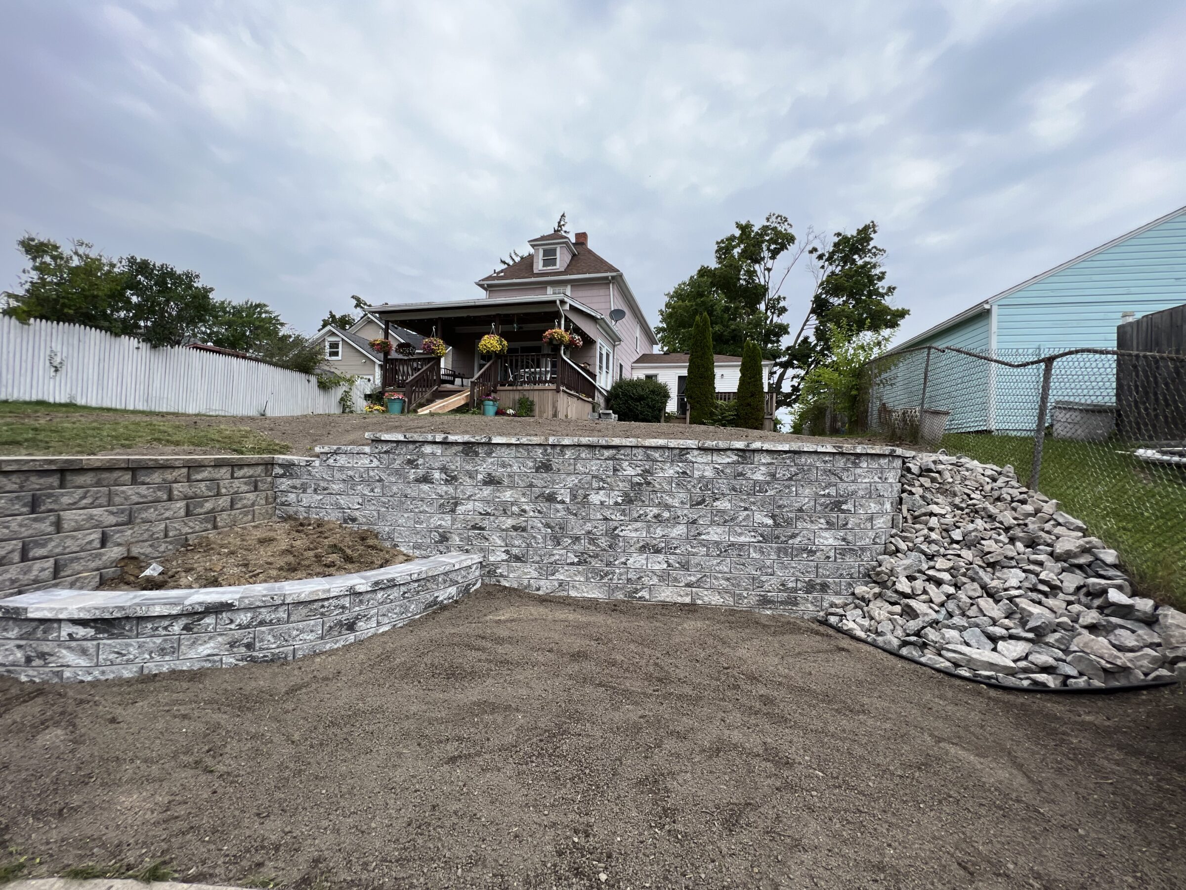 A landscaped yard with a rock retaining wall, leading to a house with a porch, surrounded by trees and a fence on a cloudy day.