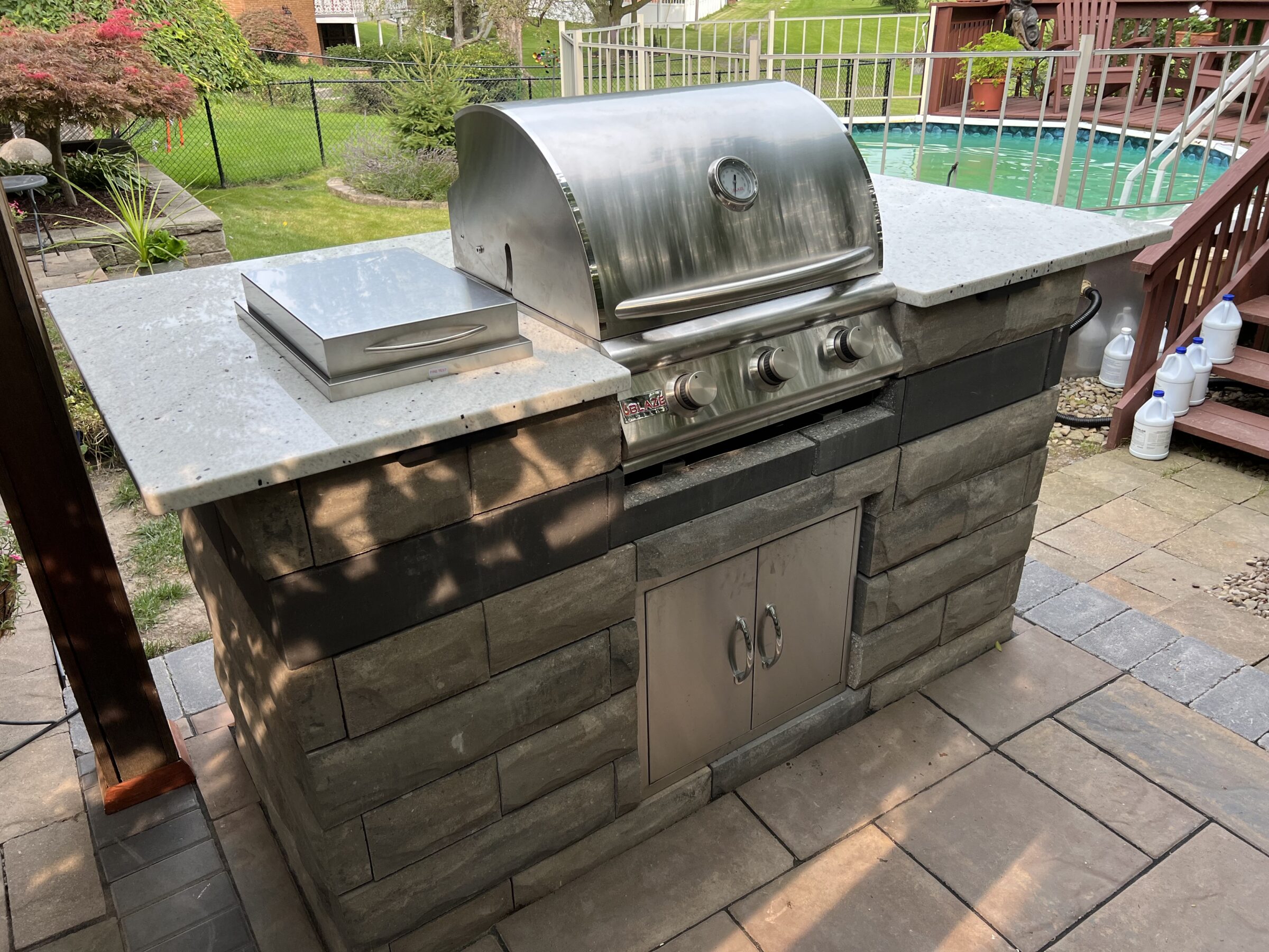 A stainless steel outdoor grill on a stone patio, surrounded by a backyard with a swimming pool and greenery in the background.