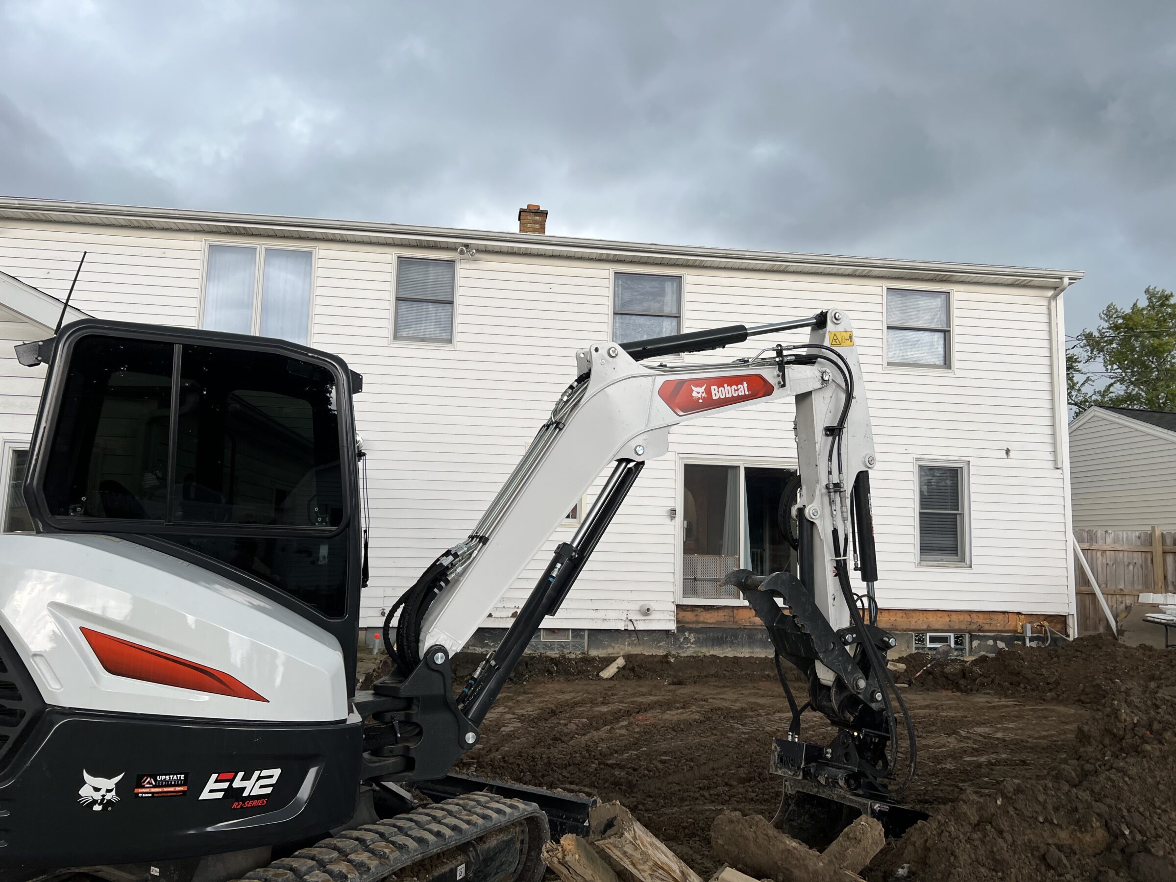 A white excavator works near a two-story house under a cloudy sky. The ground is dug up, suggesting construction or landscaping activity.