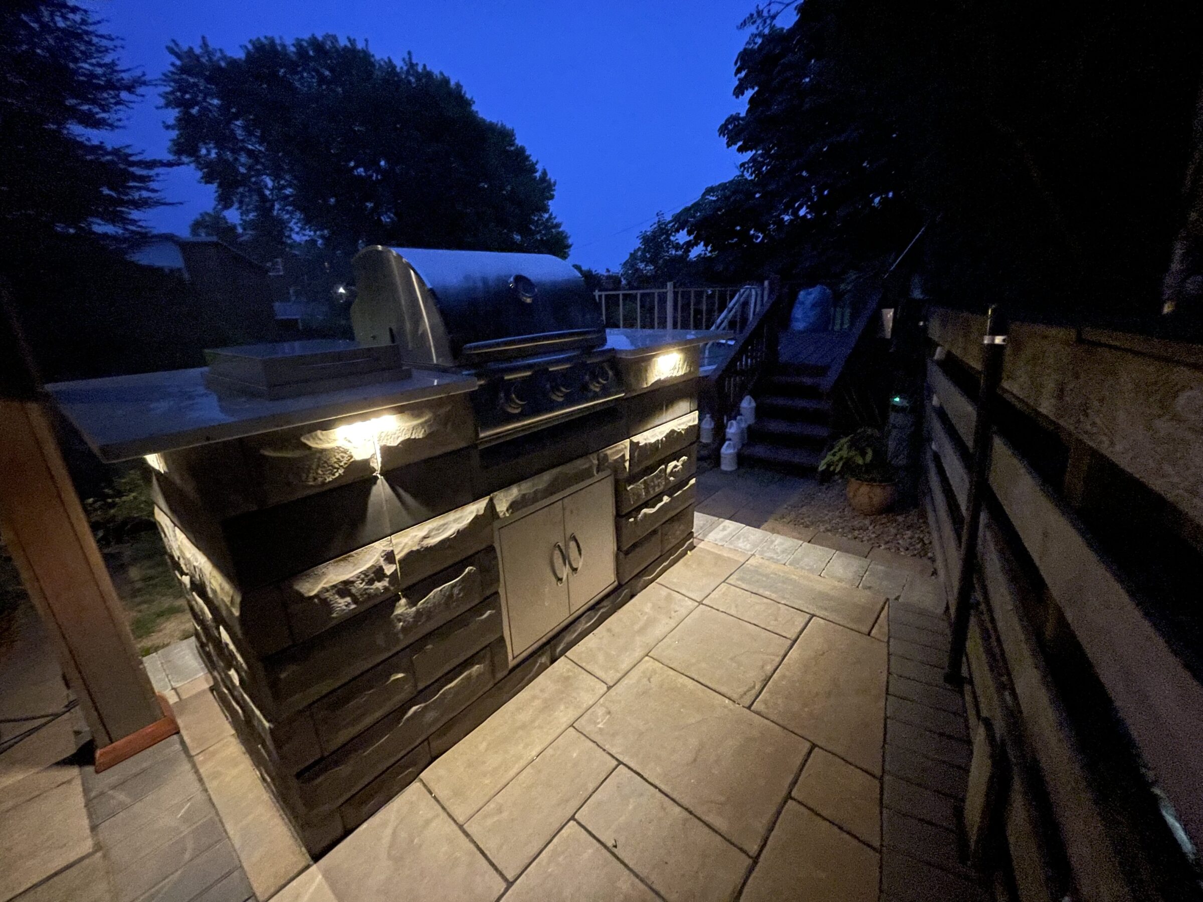 An outdoor grill area illuminated at dusk, surrounded by trees and paved stones, creates a cozy and inviting atmosphere. No people visible.