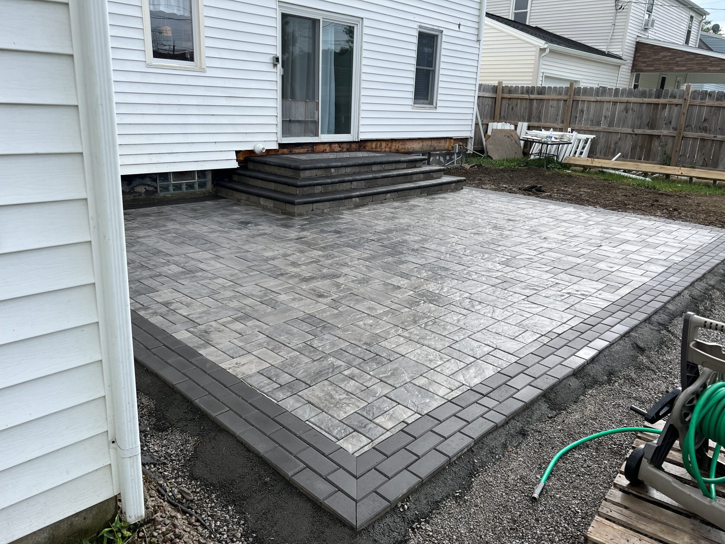 A newly constructed stone patio behind a white house with sliding doors, bordered by a wooden fence. Some garden tools are visible nearby.