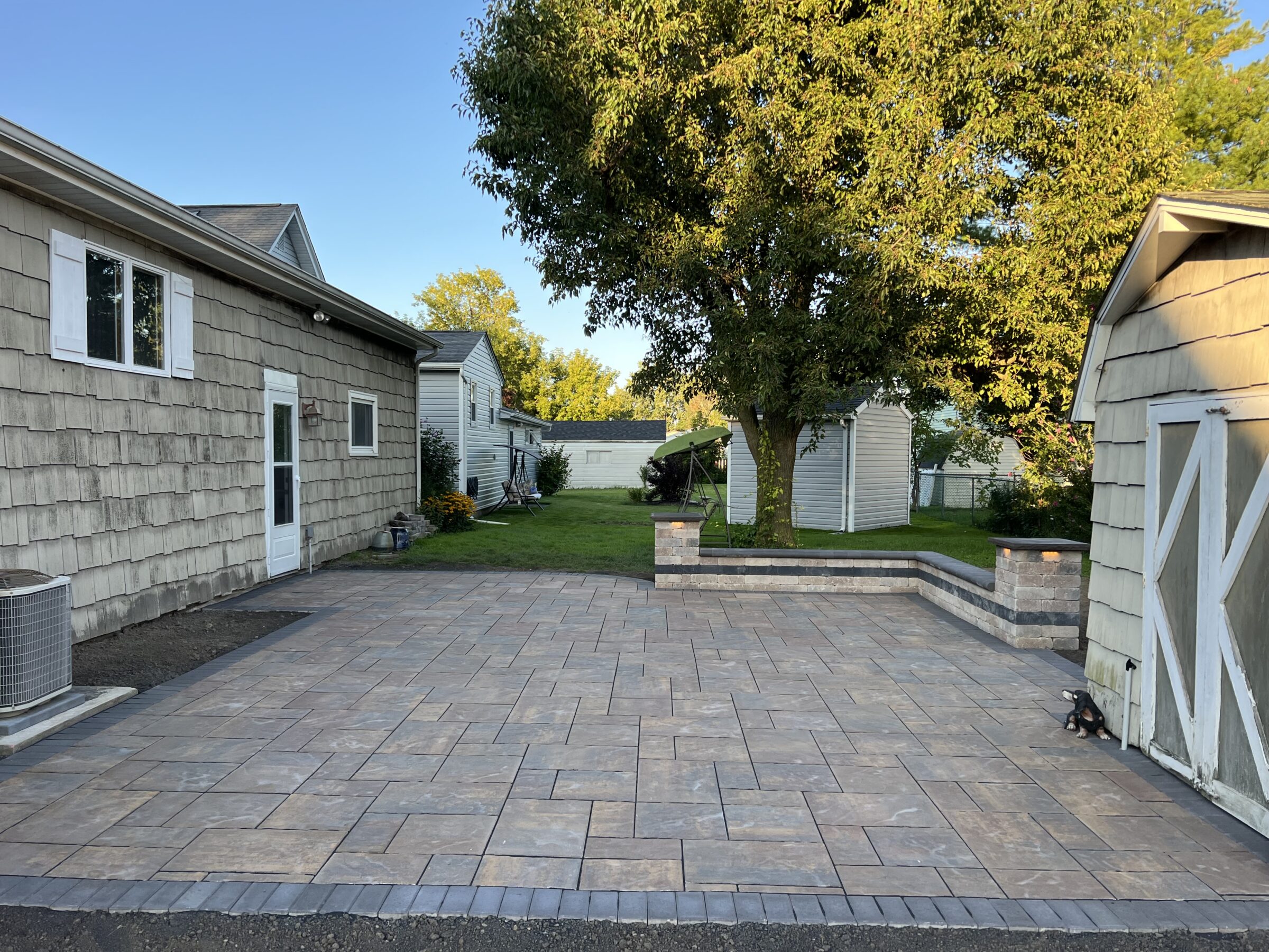 A spacious patio with stone tiles leads to a backyard, bordered by two sheds and featuring a large tree under a clear sky.