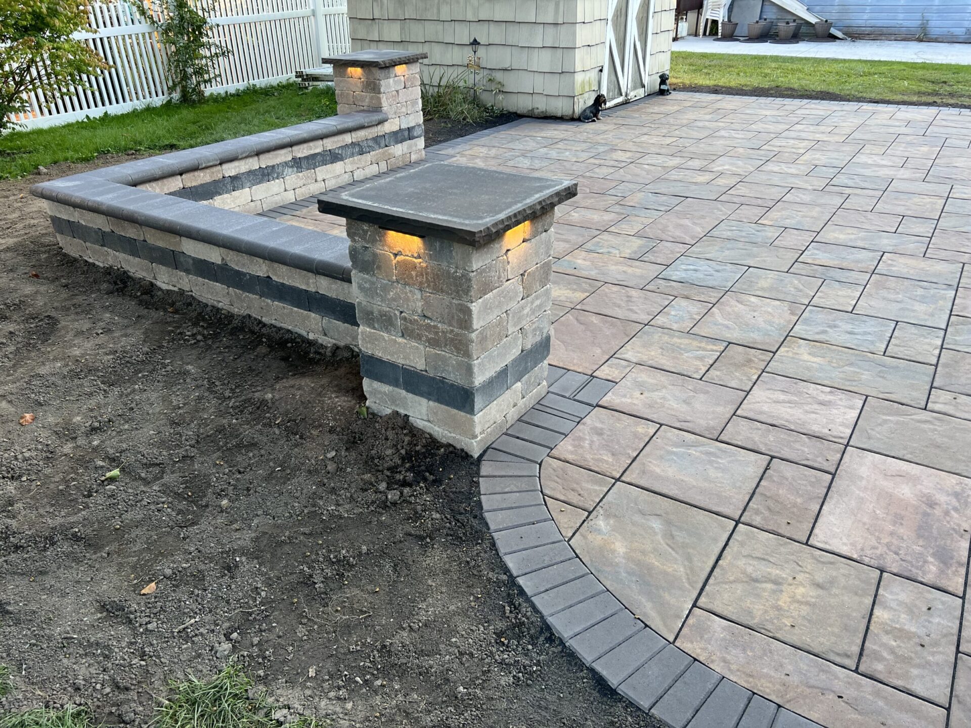 A newly paved patio with neatly arranged stone tiles and a low wall, featuring inset lighting, adjacent to a garden with fencing.