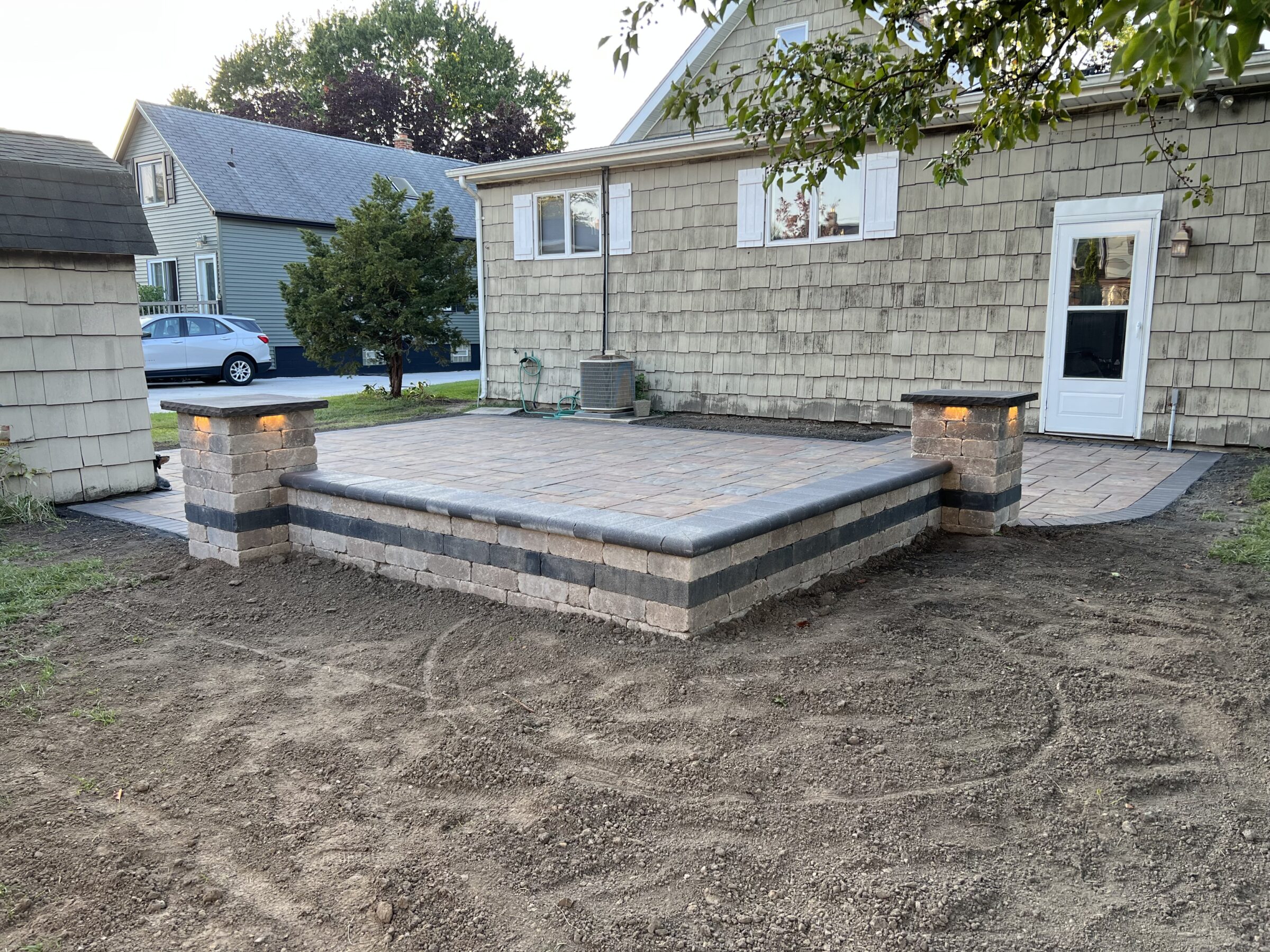 A newly constructed stone patio with illuminated pillars is attached to a house. The area around the patio is bare soil.