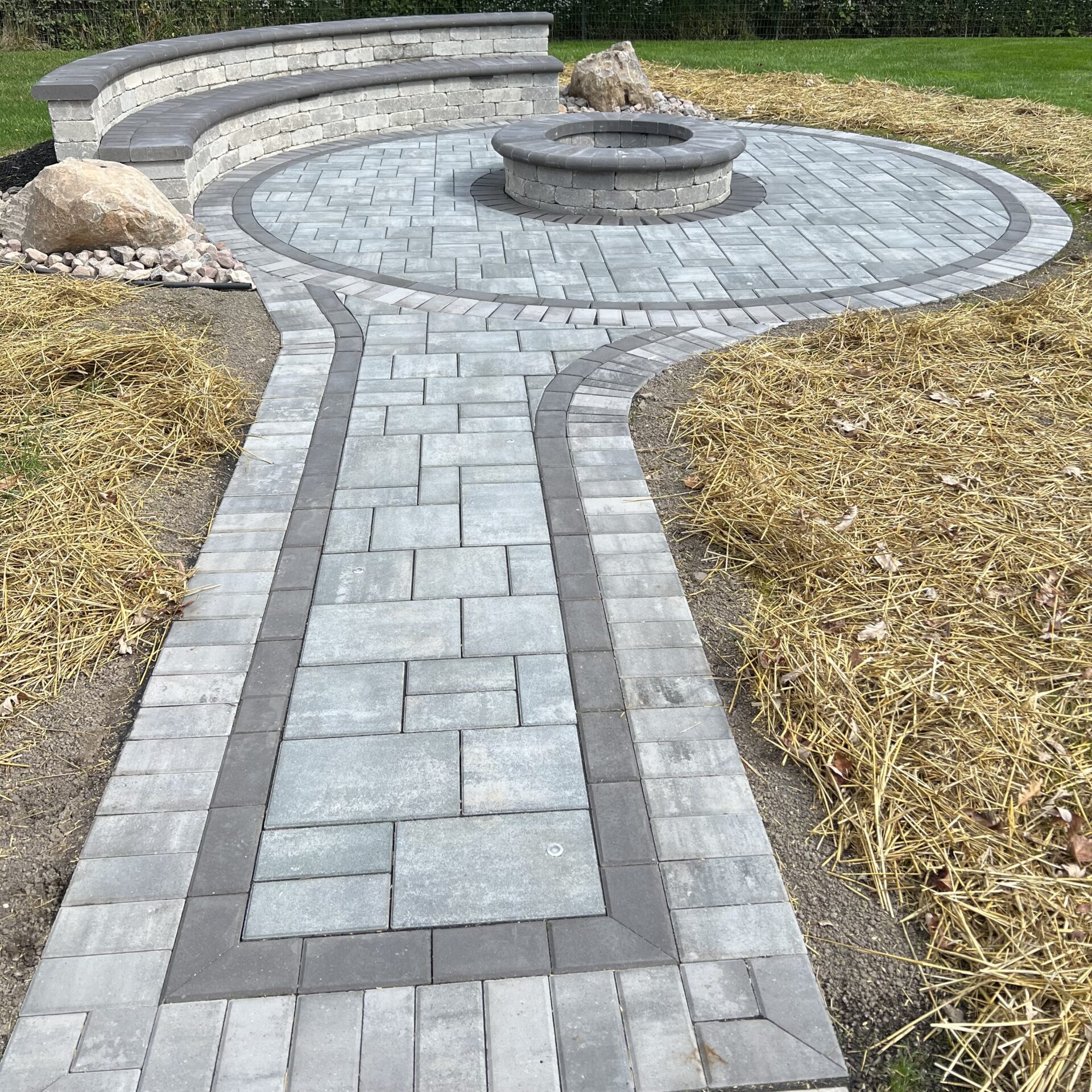 Stone patio with circular fire pit and curved bench, surrounded by straw-covered ground. No recognizable landmarks or historical buildings present.