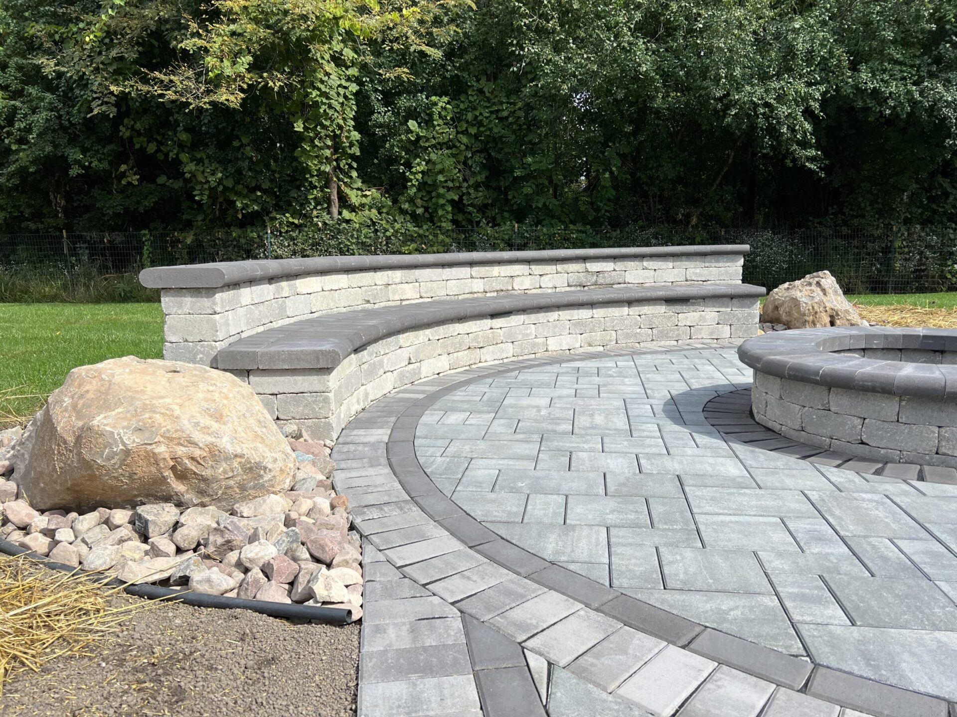 Curved stone bench and fire pit on a paved patio, surrounded by rocks and greenery, under a clear sky.