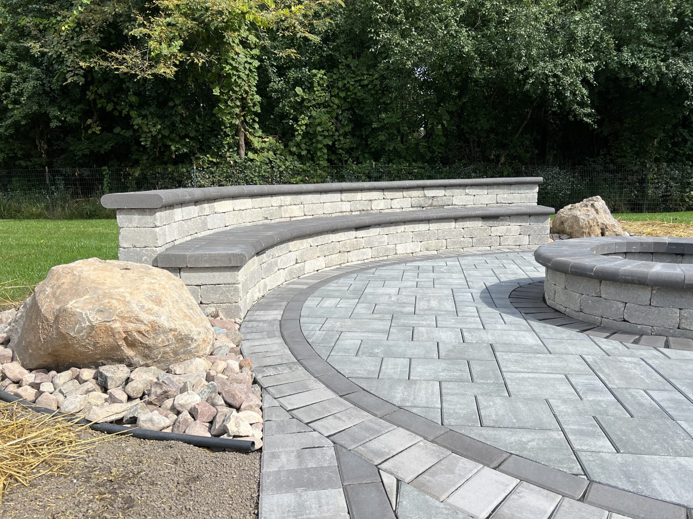 Curved stone bench and pathway with boulders, surrounded by greenery. Features a circular stone seating area, suggesting a backyard patio setting.