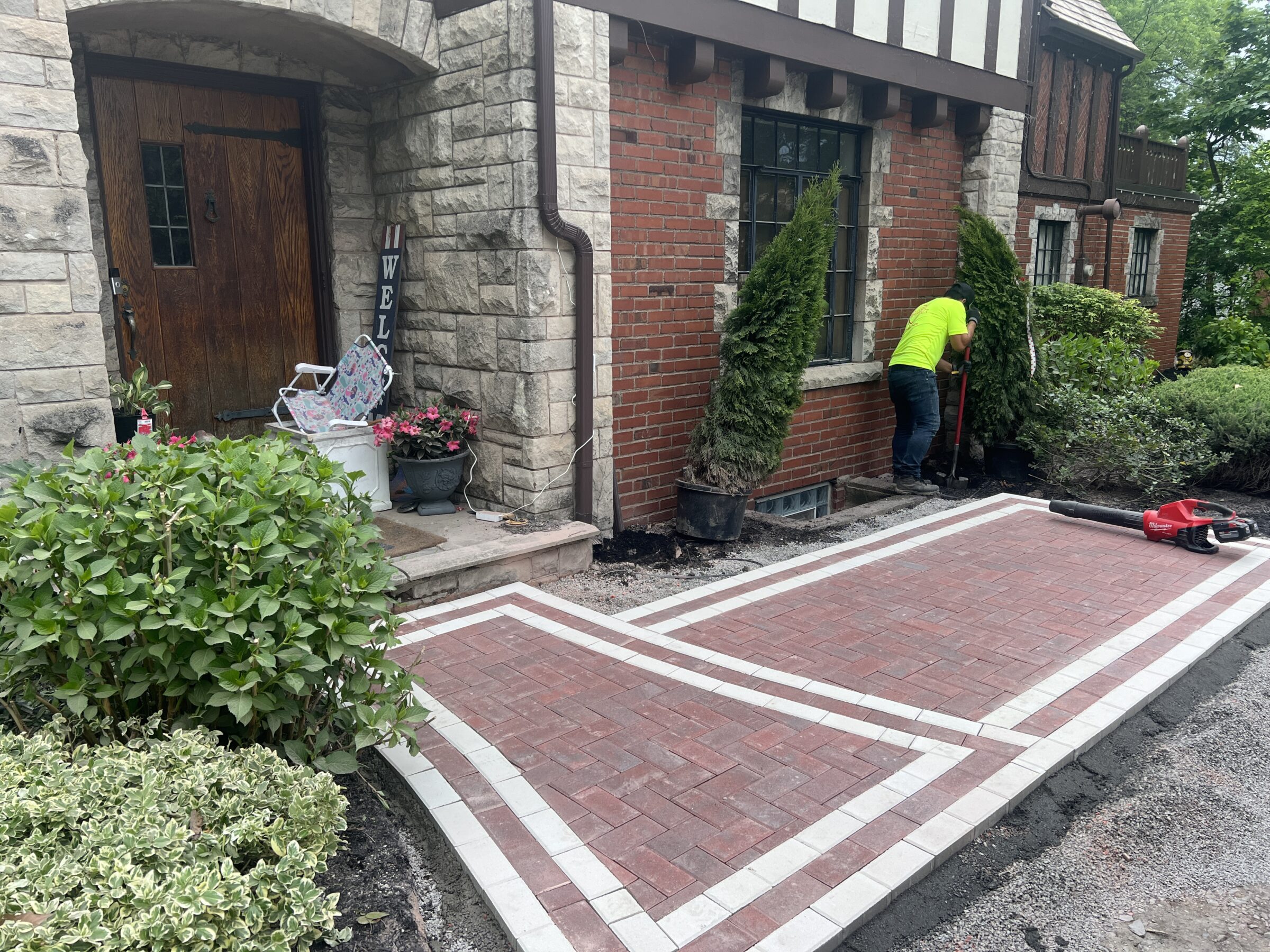 A person in a bright shirt works on landscaping outside a brick and stone house. Decorative pavers form a new pathway.