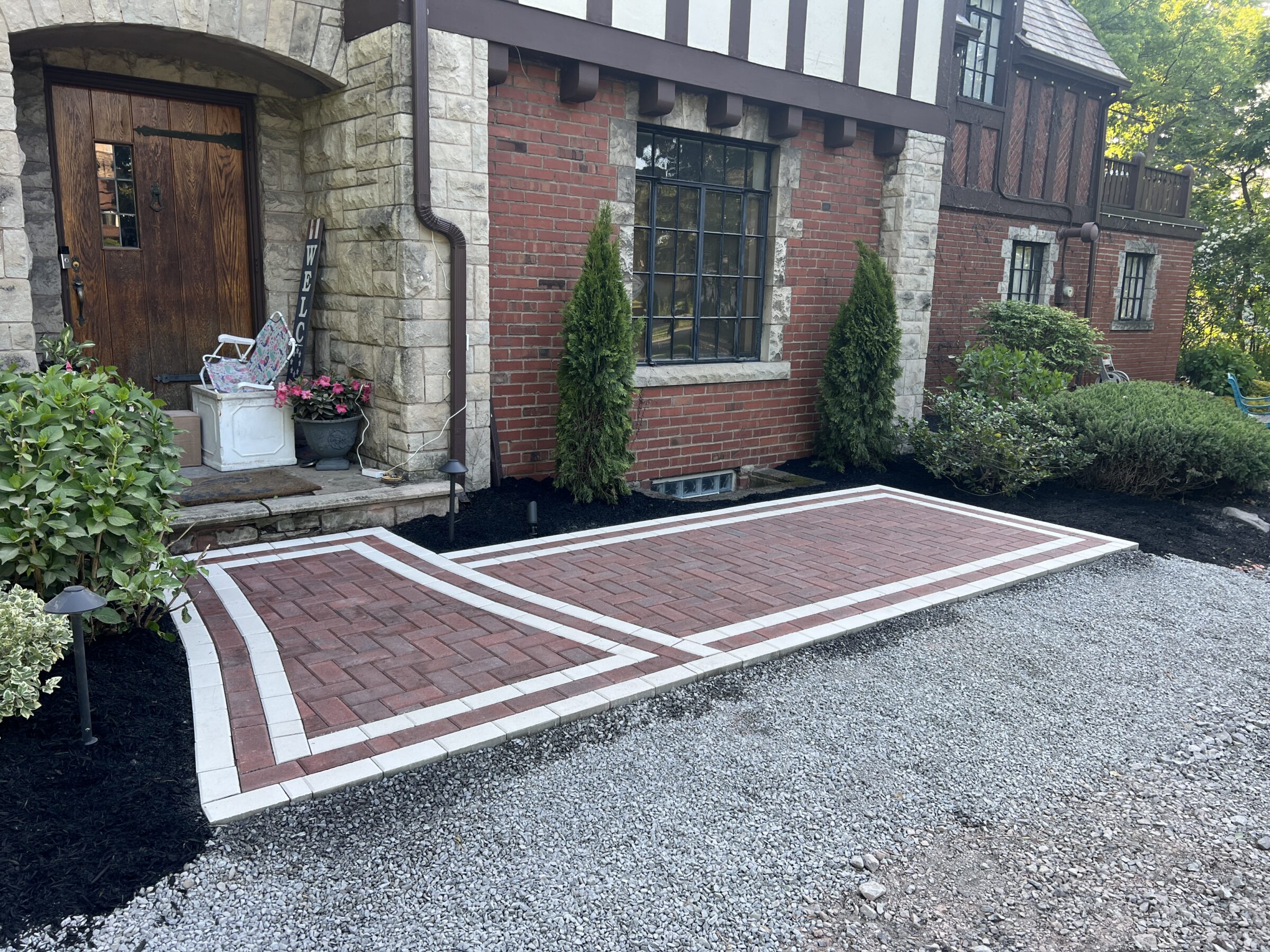 Brick path with white trim leads to a wooden door on a Tudor-style house. Surrounding greenery and gravel define the landscaped entrance.