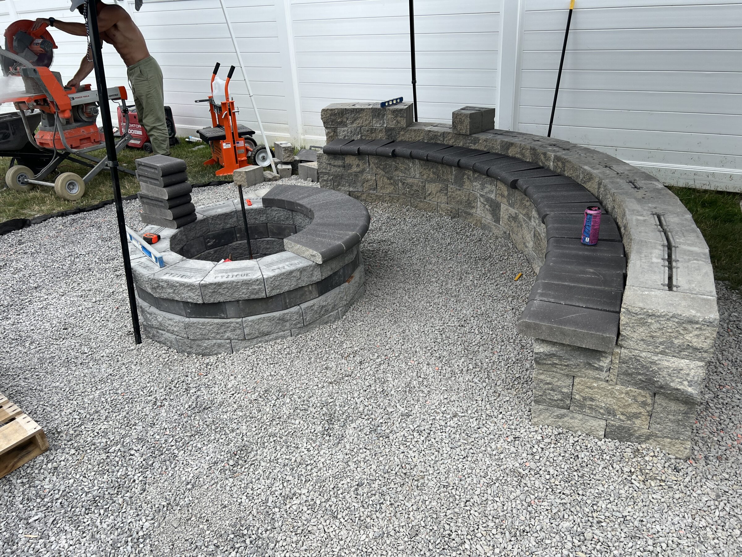 A circular stone fire pit and curved bench under construction on gravel, with a person working nearby, and tools visible.