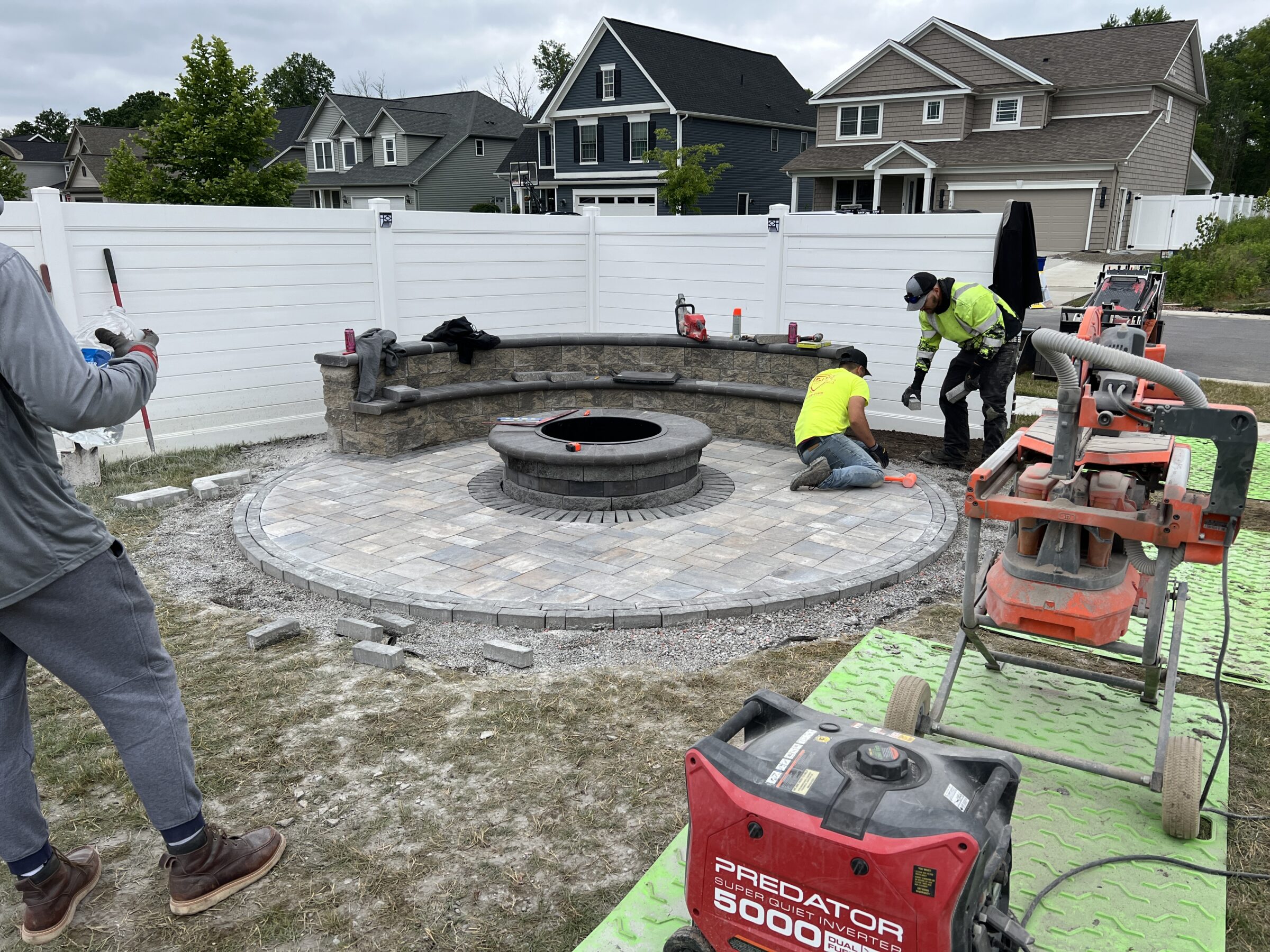 Two people work on a circular stone patio with fire pit. Residential homes and construction equipment are visible nearby.