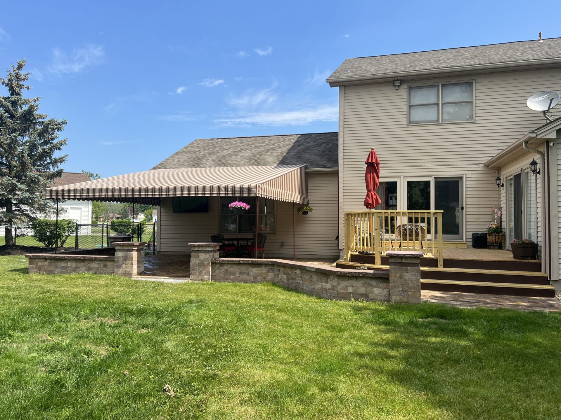 A spacious backyard with a patio, covered seating area, and deck adorned with a red umbrella. Lush grass and trees surround the house.