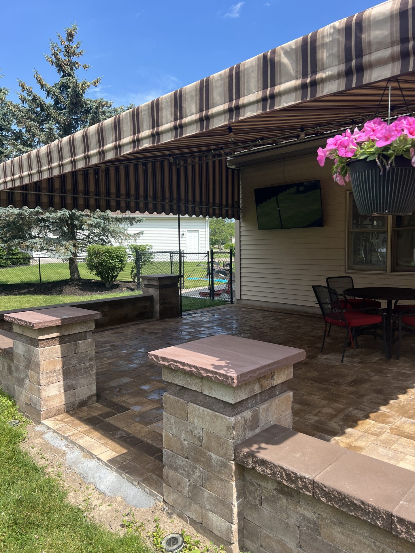 A covered patio with a striped awning features outdoor seating, a television, and potted flowers, surrounded by a grassy yard and trees.