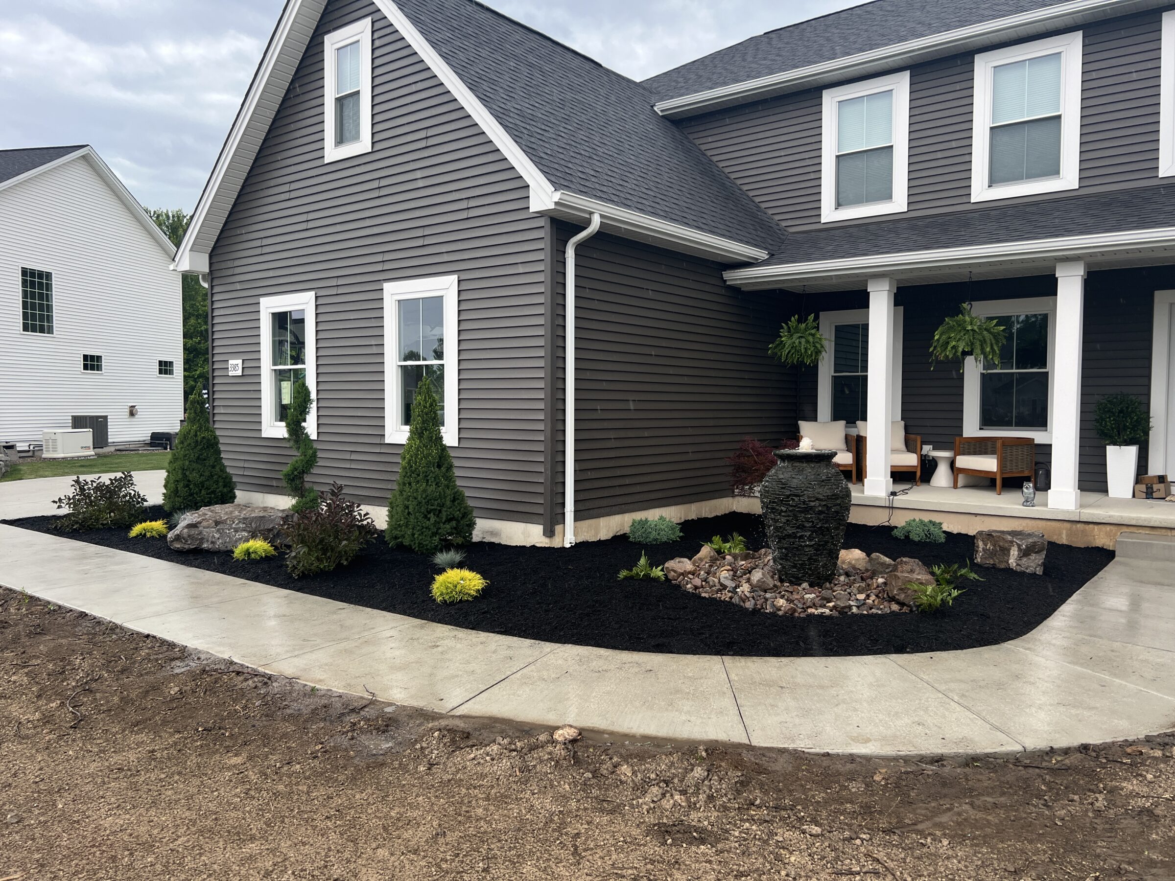 A modern gray house with a landscaped front yard, featuring mulch beds, shrubs, and a stone fountain beside a concrete walkway.