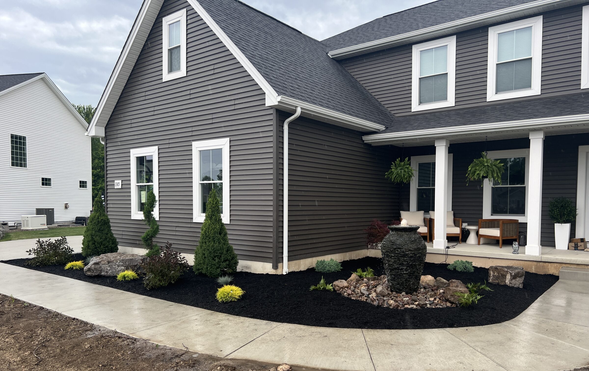Modern gray house with a landscaped front yard, featuring shrubs and a water fountain. Porch with seating. Nearby, another white house is visible.