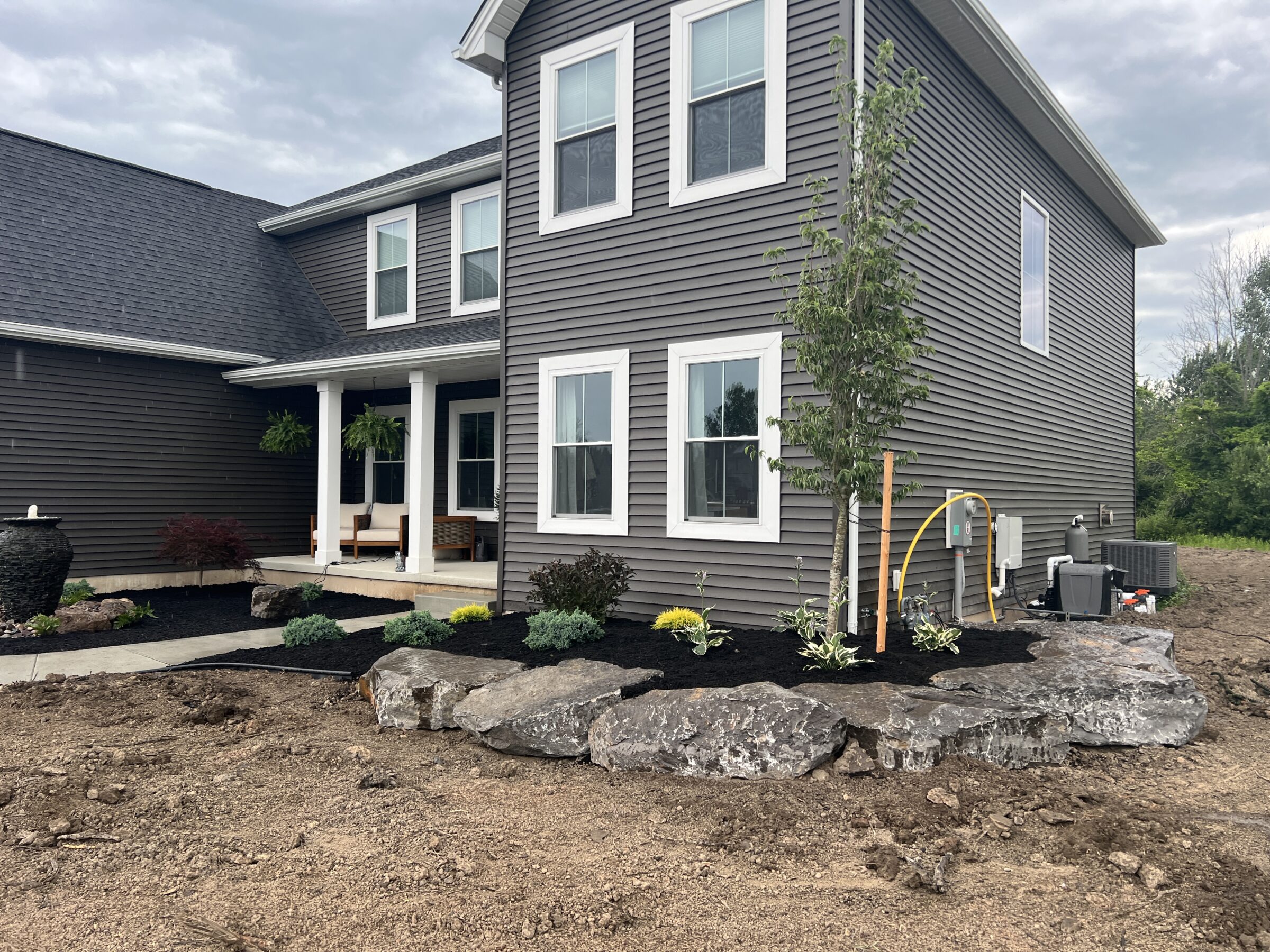 A modern gray two-story house with a landscaped garden, featuring rocks and small plants, surrounded by bare soil and a cloudy sky.