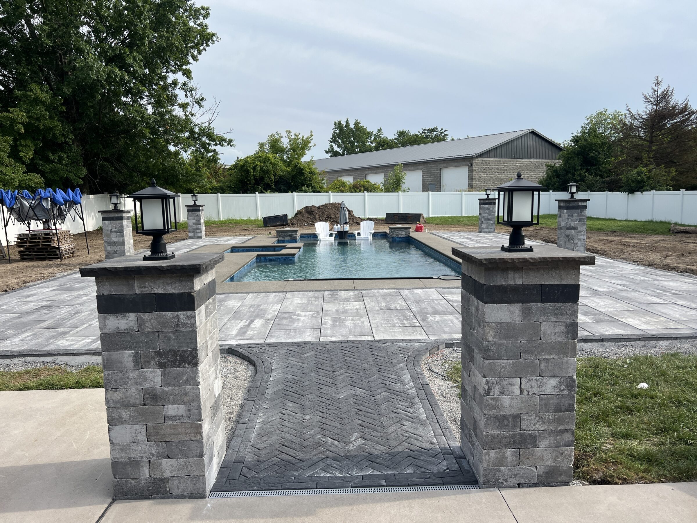 A modern outdoor space with a rectangular pool, surrounded by stone tiles and lantern-topped pillars. White fence and trees in the background.