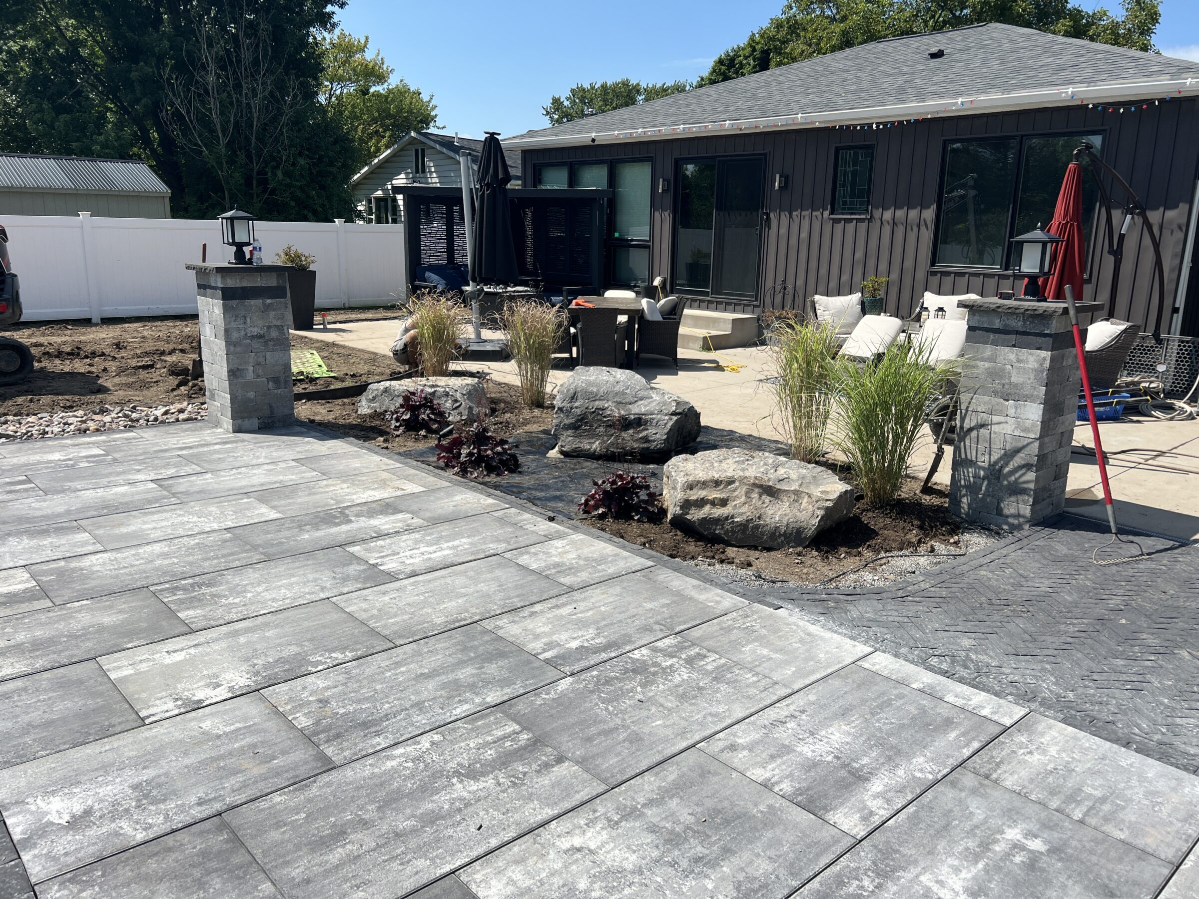 A modern backyard features a paved patio, decorative stones, plants, and seating. A house and fence provide a cozy, private atmosphere.