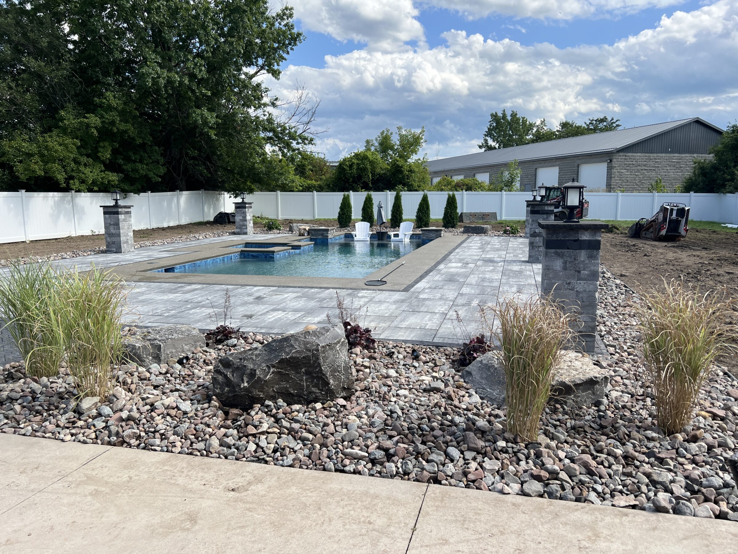 A landscaped backyard features a modern swimming pool with stone decking, surrounded by white fencing and trees, under a partly cloudy sky.