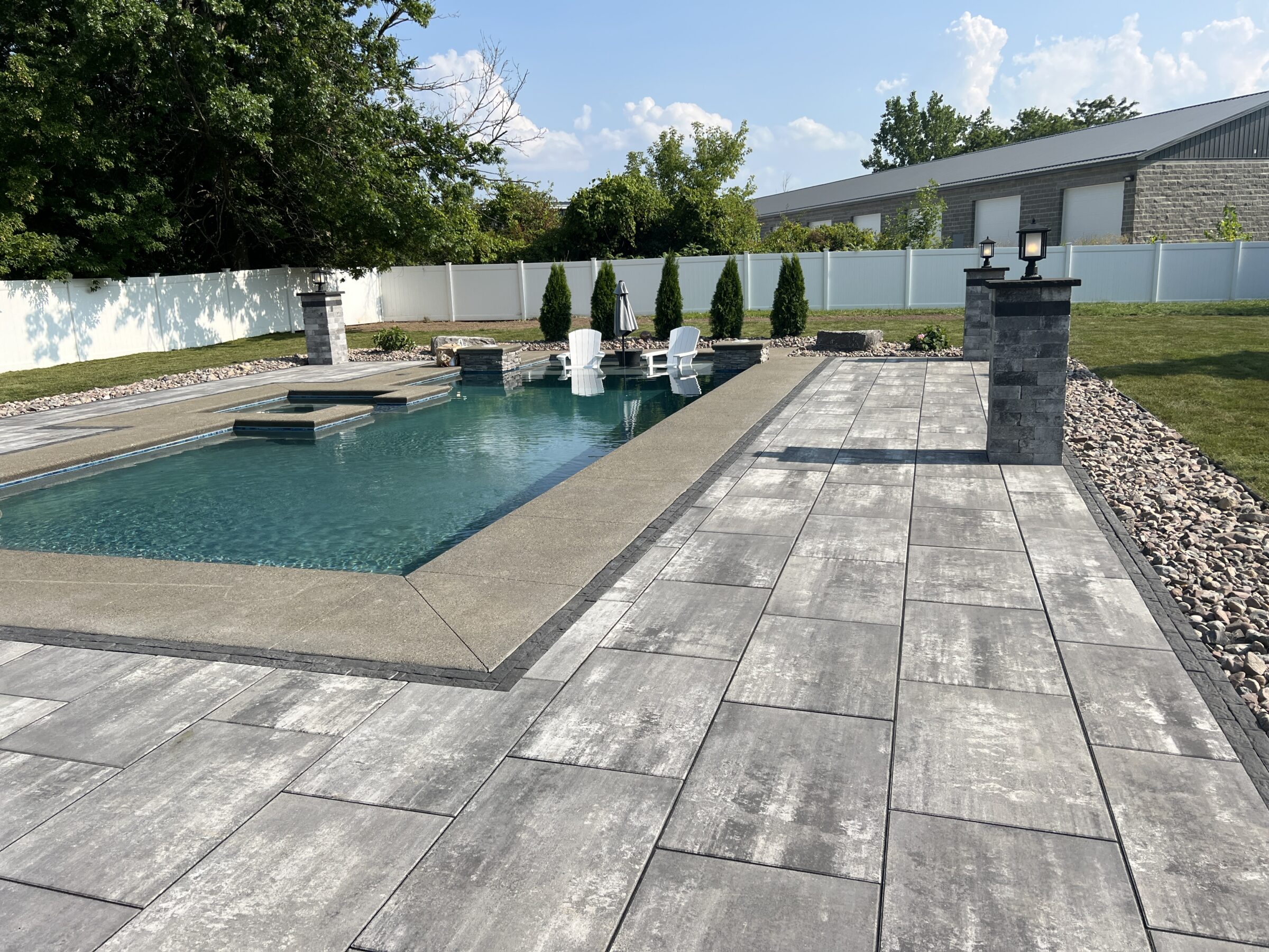 A modern outdoor pool with stone patio, surrounded by greenery and a privacy fence. Two chairs are placed near the water's edge.