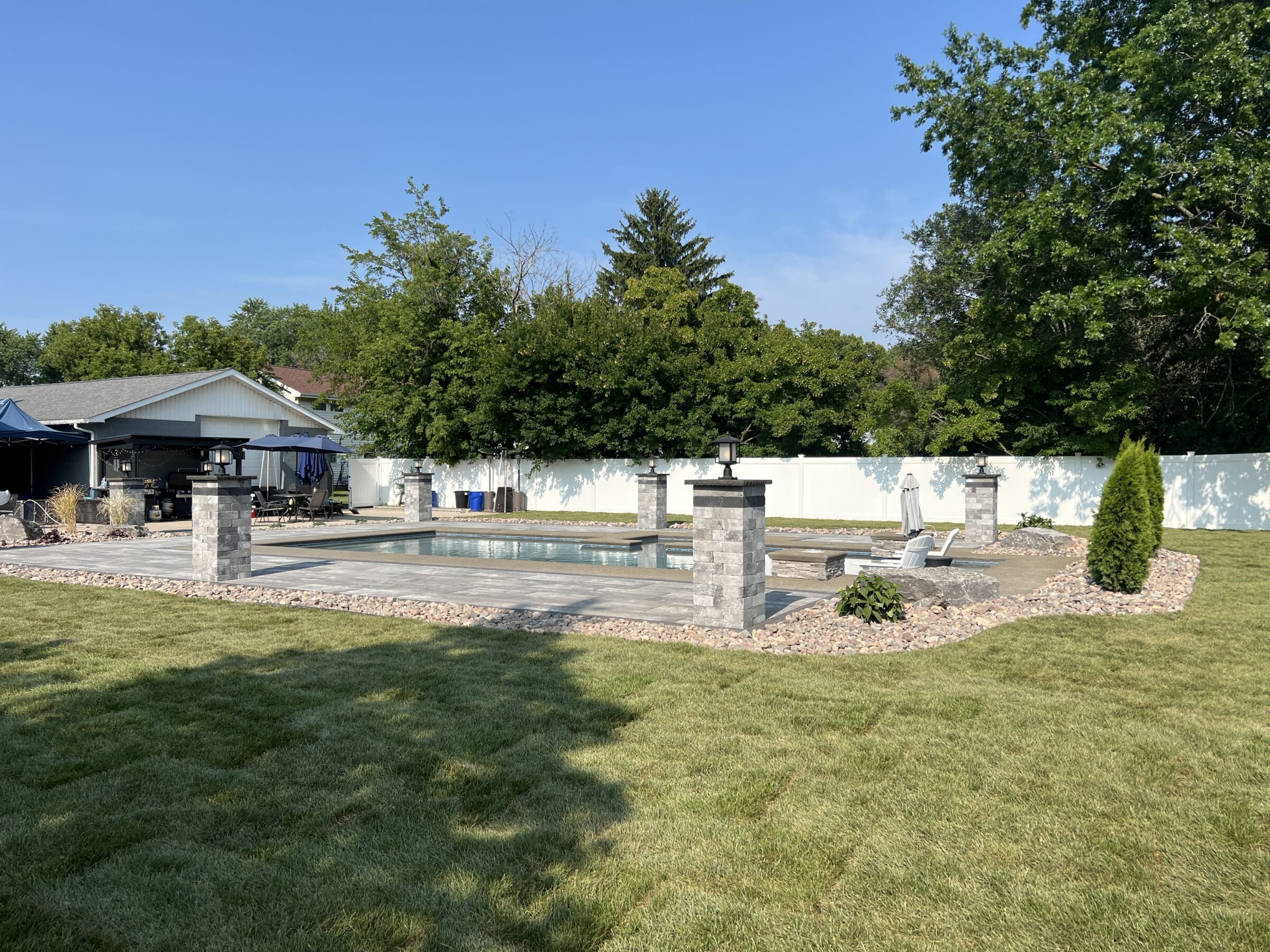 A backyard scene features a rectangular swimming pool with stone pillars, surrounded by grass and trees, under a clear blue sky.