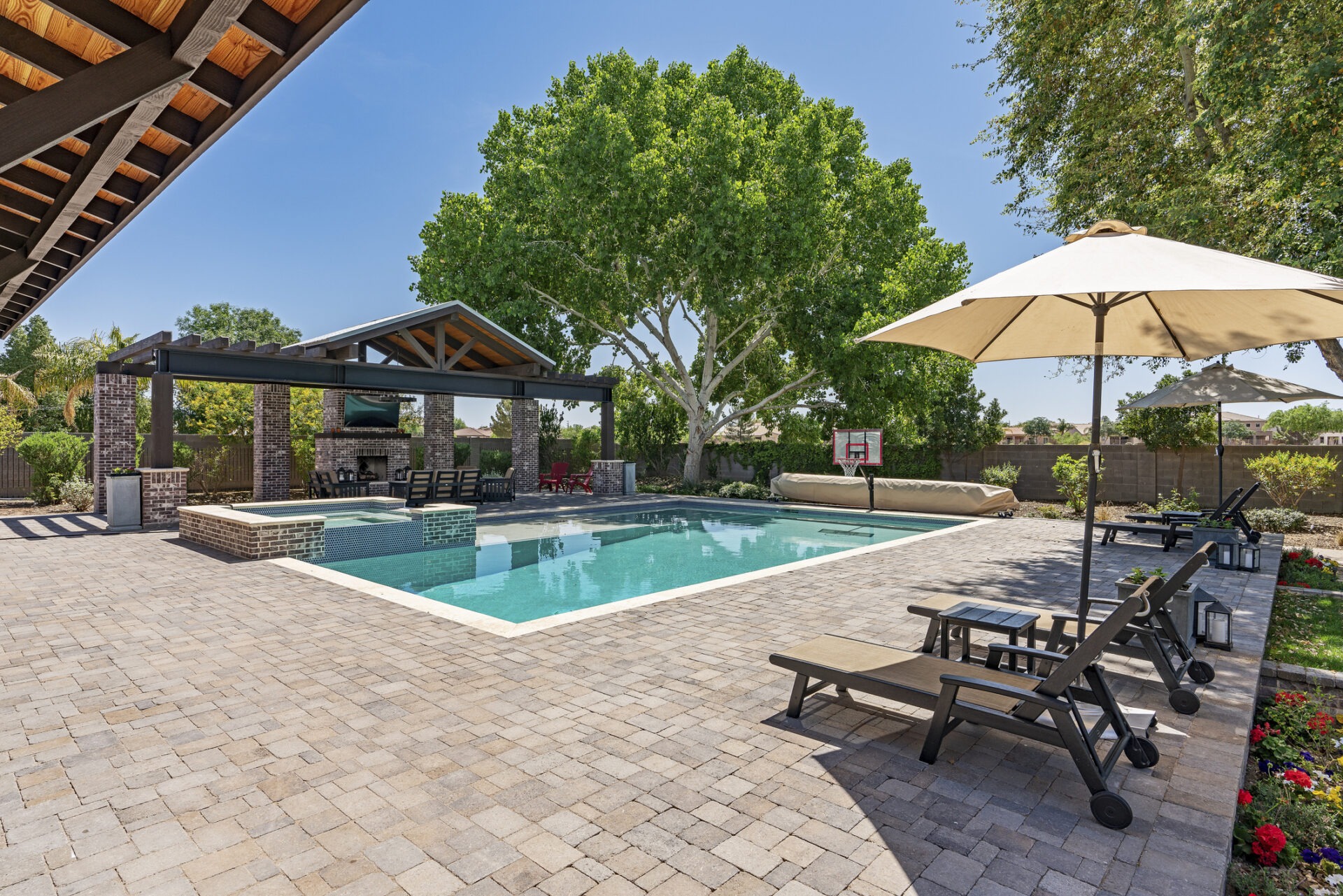 A backyard features a swimming pool, lounge chairs, a large umbrella, a pergola with seating, and lush trees under a clear blue sky.