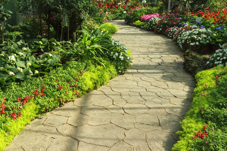 A winding stone path bordered by vibrant flowers and lush greenery under soft sunlight, creating a serene and colorful garden scene.