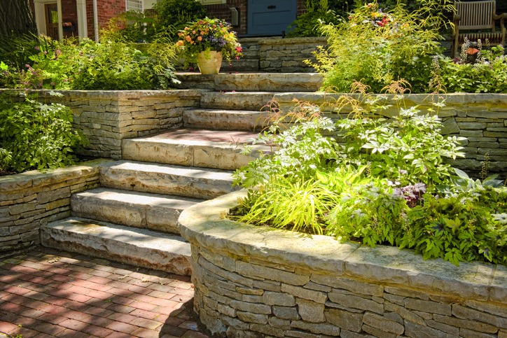 Stone steps with lush green plants and flowers lead to a brick house entrance, creating a welcoming and serene garden atmosphere.