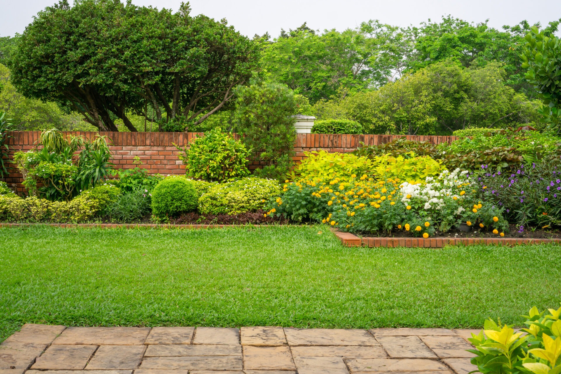 A lush garden features vibrant flowers, neatly trimmed bushes, and a brick wall, with a paved stone path in the foreground.