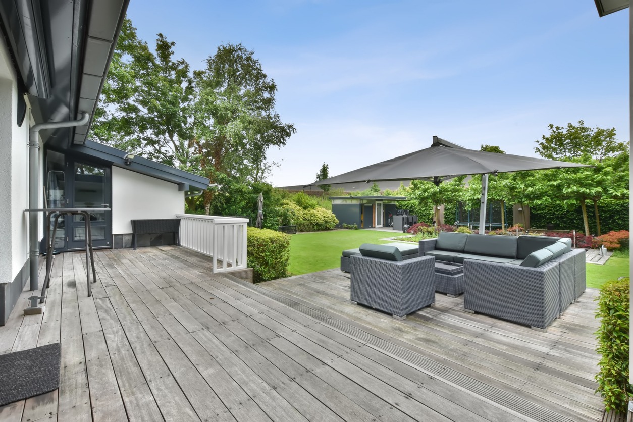 Spacious backyard features a wooden deck, modern outdoor furniture, and large umbrella. Lush greenery and trees surround the well-maintained lawn and patio.