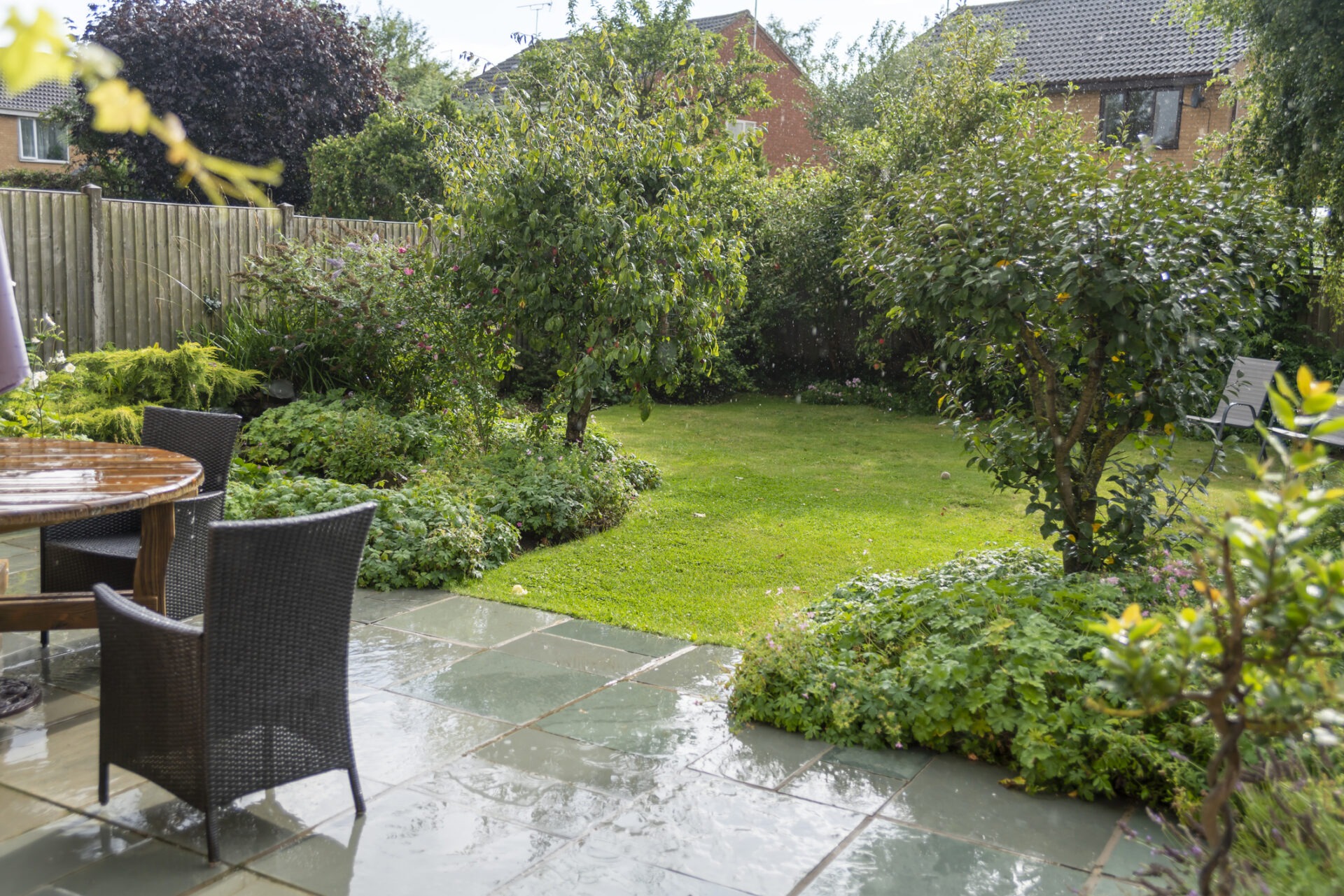 A cozy garden with lush greenery, patio furniture, and wet paving, surrounded by a wooden fence and houses in the background.