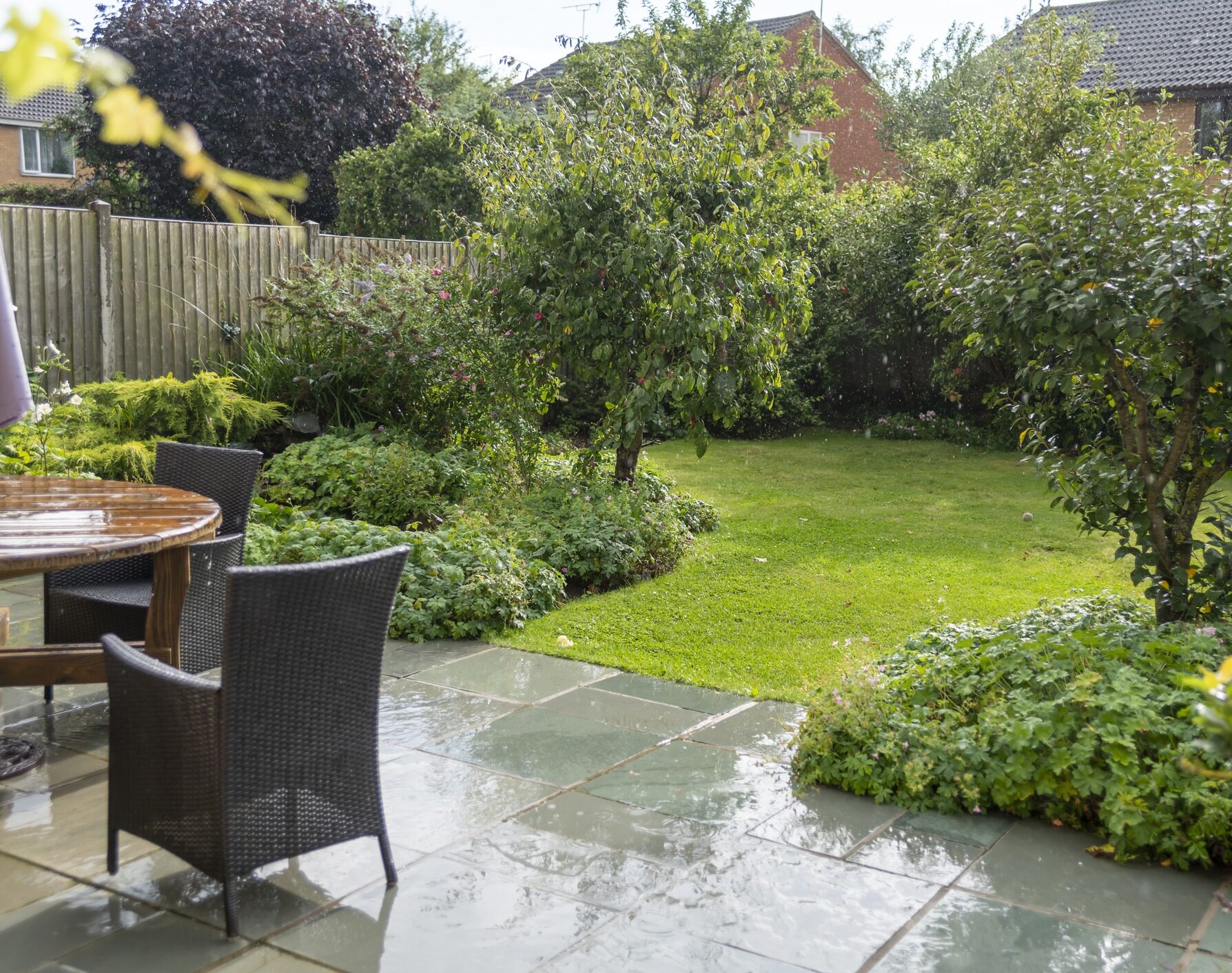 A cozy garden with lush greenery, patio furniture, and wet paving, surrounded by a wooden fence and houses in the background.