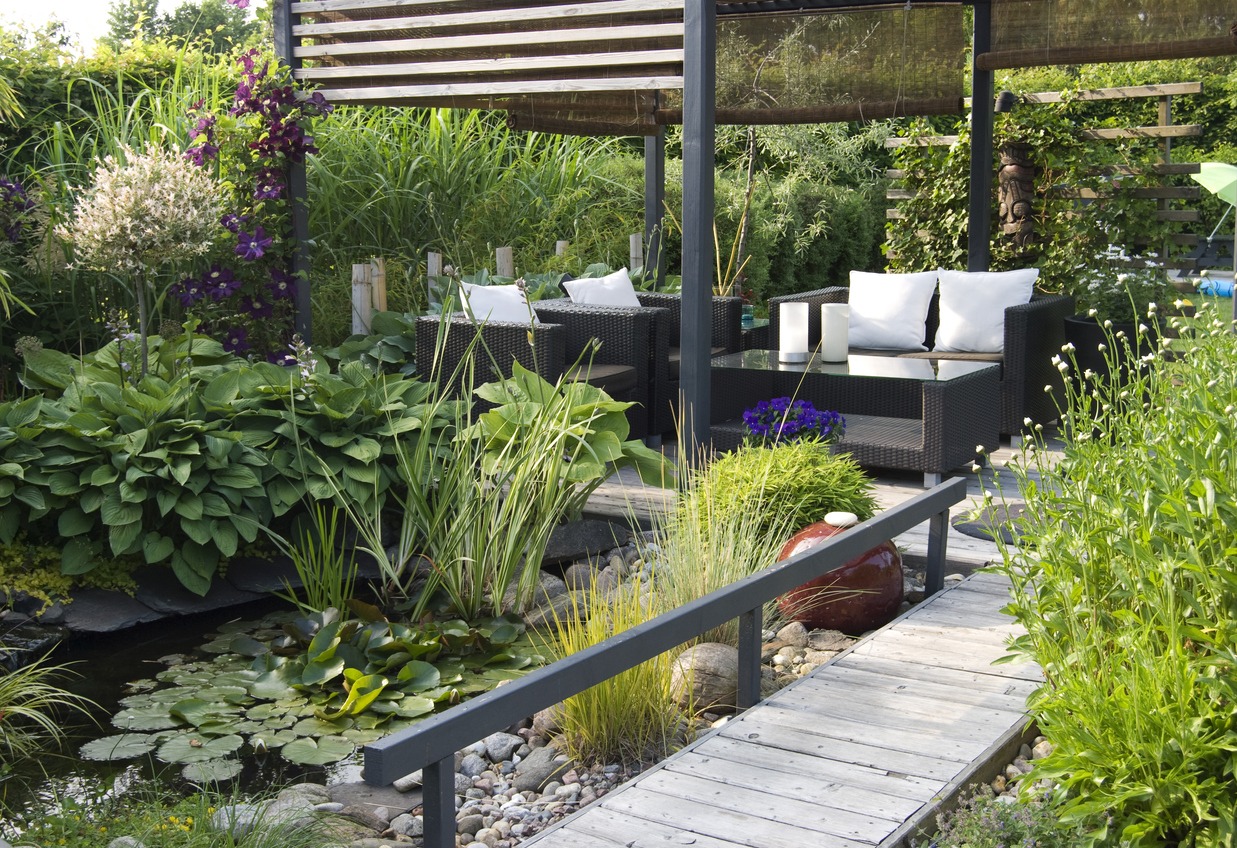 A serene garden with lush greenery, a small pond, wooden walkway, and a shaded seating area featuring modern outdoor furniture.