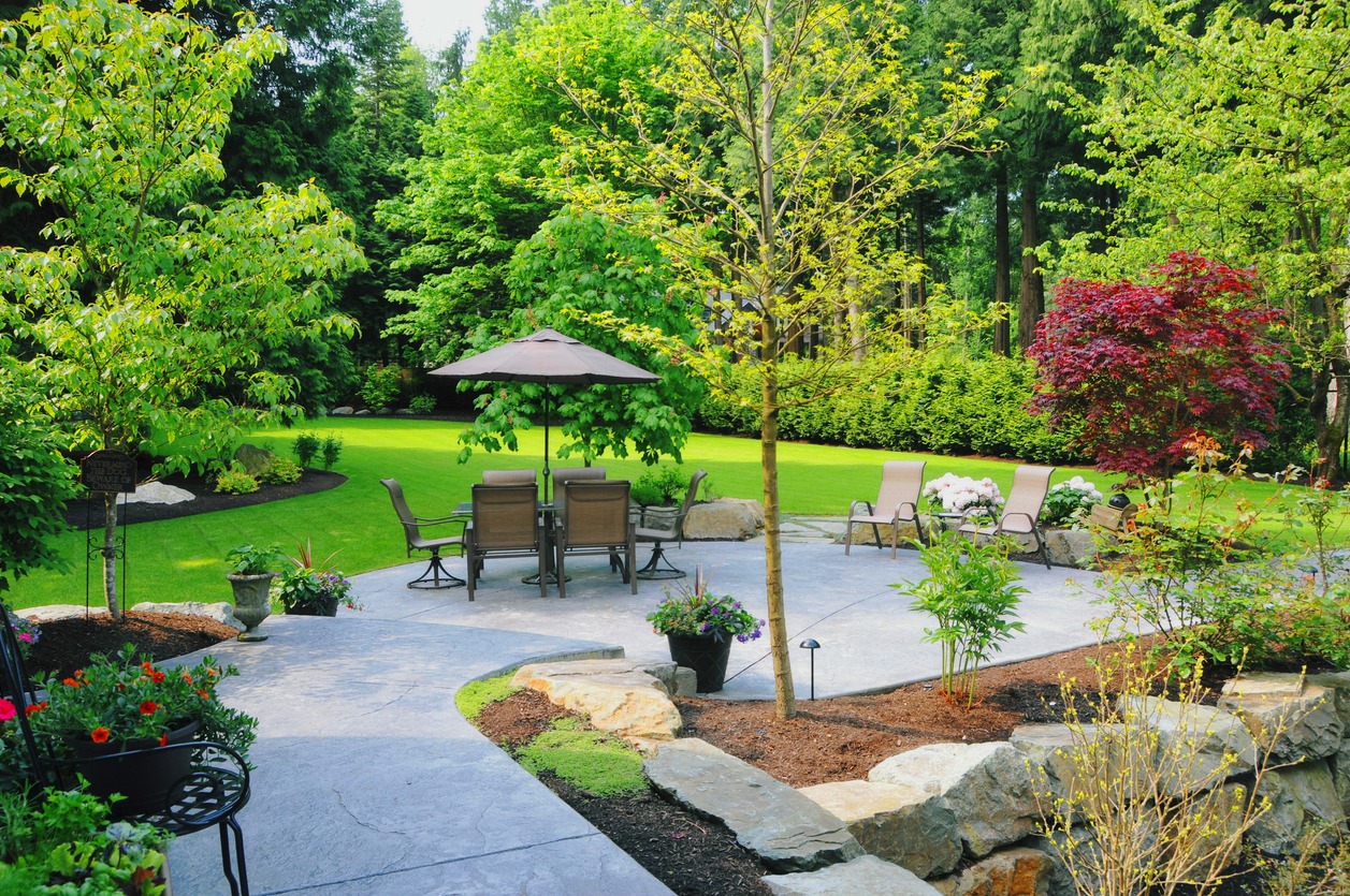 A lush garden features a patio with chairs and umbrella, surrounded by vibrant greenery, trees, and colorful plants on a sunny day.