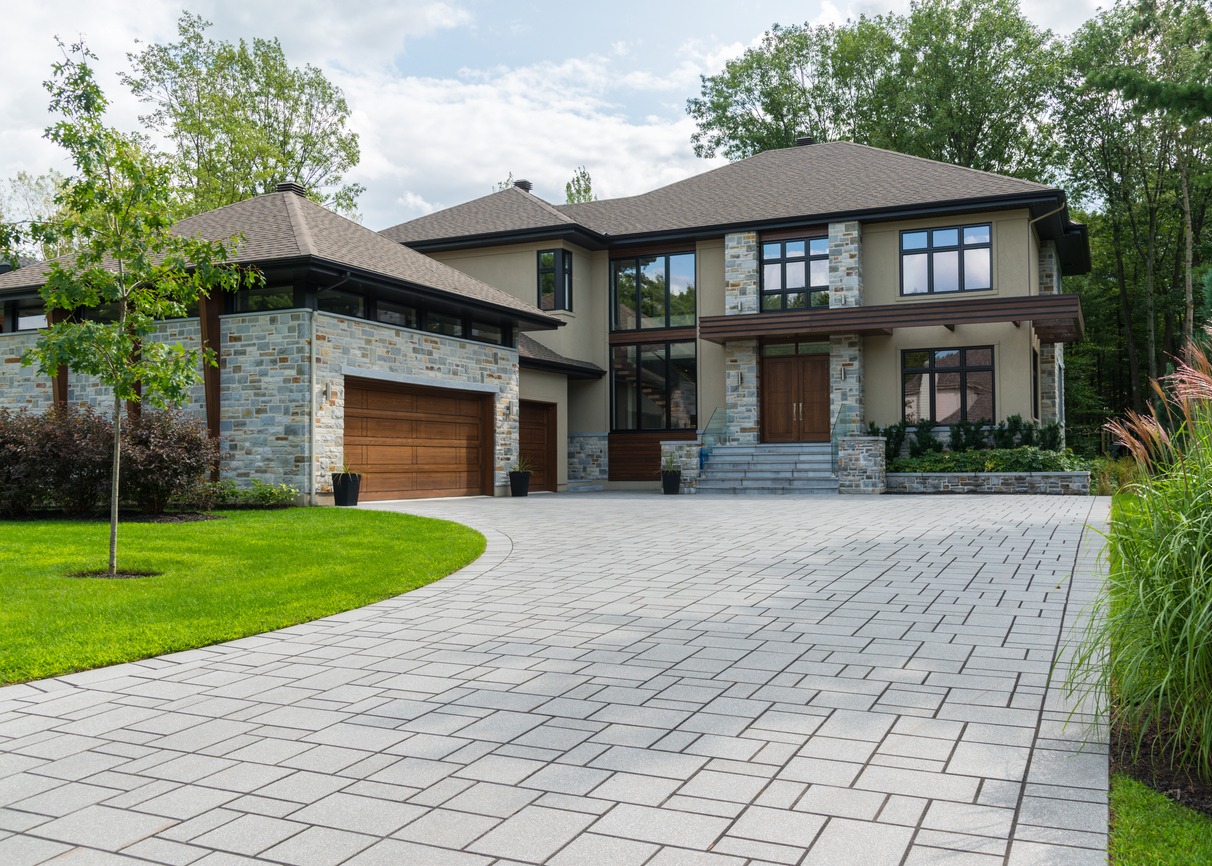 Modern two-story house with stone and wood facade, large windows, and spacious driveway, surrounded by lush greenery and landscaped lawn under a partly cloudy sky.