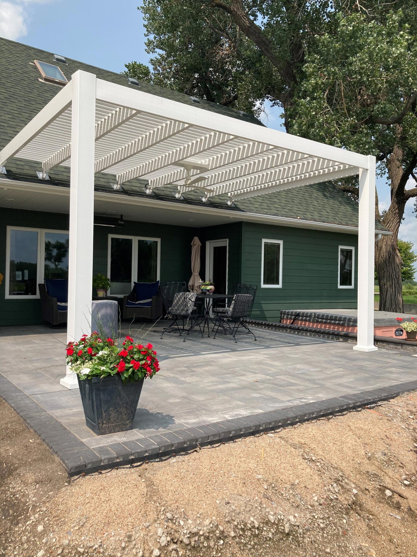 A modern green house with a covered patio, outdoor seating, and a flower pot. Surrounded by trees and natural landscaping.