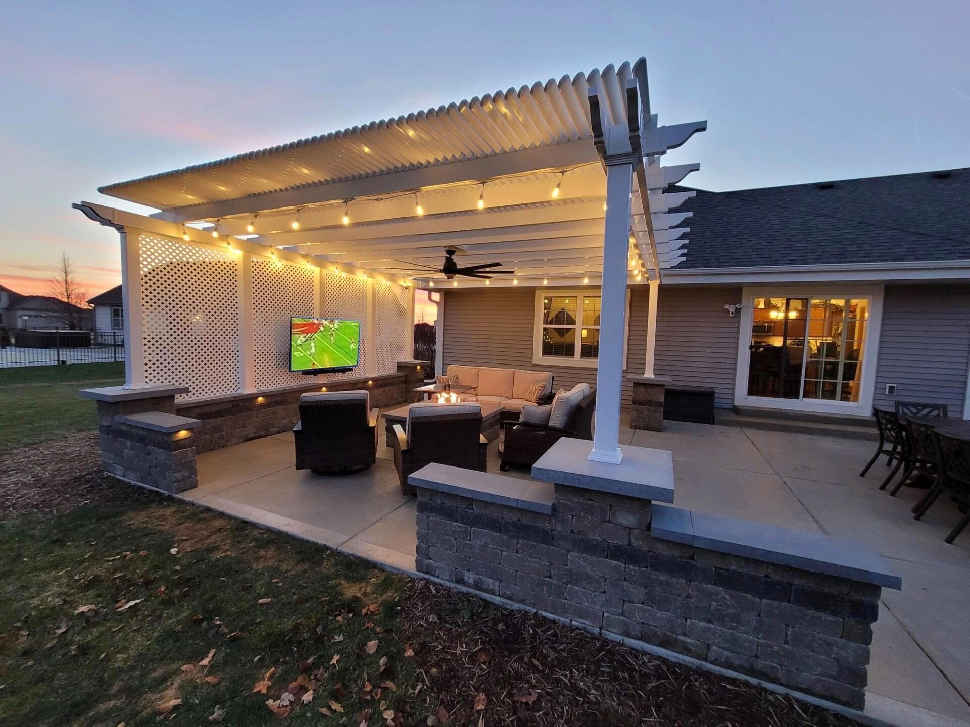 A backyard patio with pergola, string lights, and outdoor furniture. A TV is on the wall, displaying a soccer match as the sun sets.